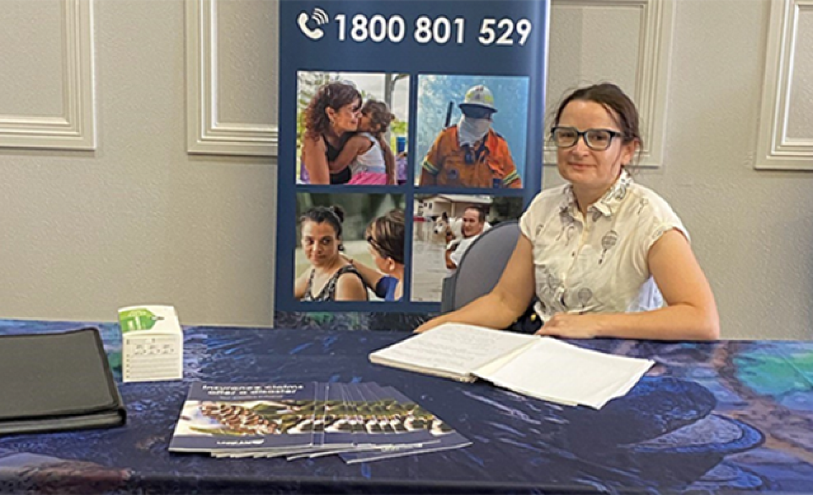 Emma sits at a table. There is a banner behind her with the phone number for the Disaster Response Legal Service on it.  banner image