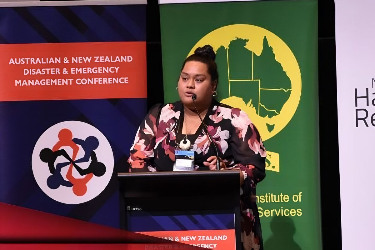 Ma'ata stands behind a podium. She is mid-speech. Behind her are brightly coloured conference banners, including one that says Australian and New Zealand Disaster and Emergency Management Conference