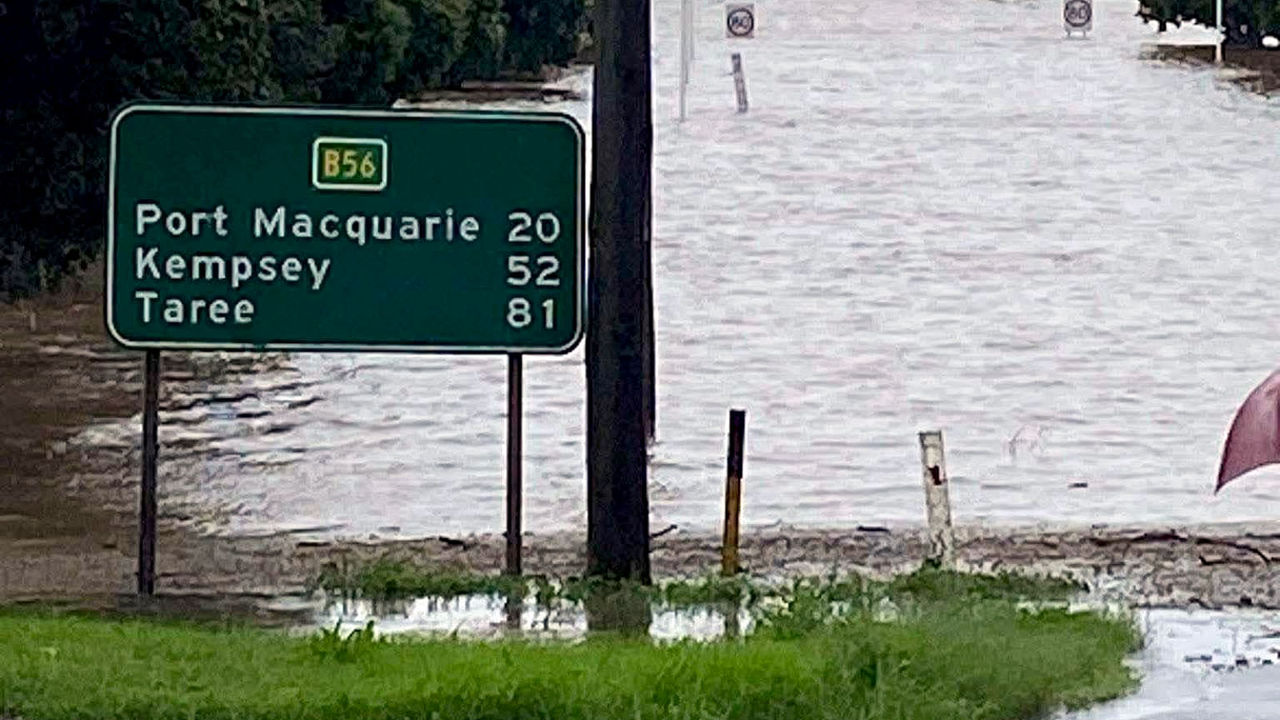 A green road sign showing distances in kilometres to Port Macquarie, Kempsey and Taree. The entire area behind the sign is flooded with water.