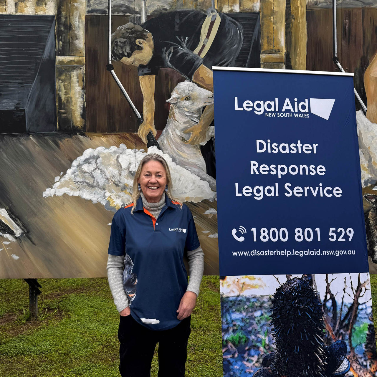 DRLS solicitor Anne Murphy standing alongside a DRLS promotion banner, with a mural sheep shearing mural behind her.