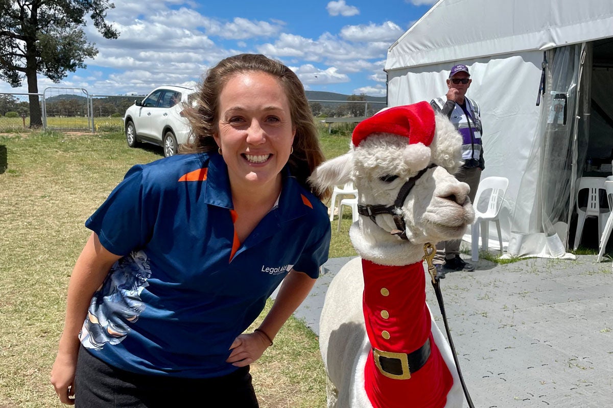 A smiling woman wearing a Legal Aid NSW tshirt stands next to an alpaca. The alpaca is dressed in a santa costume.