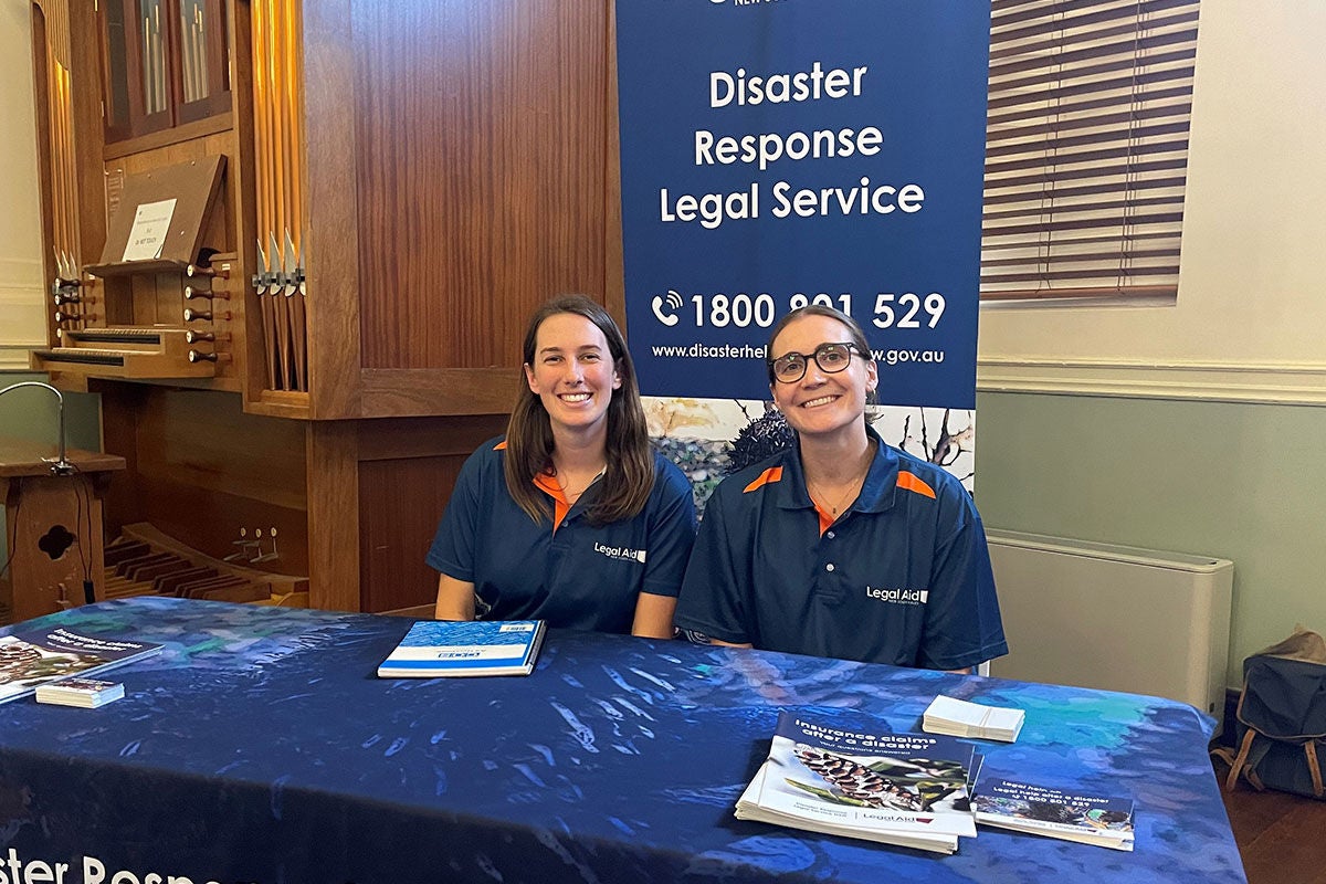 Two DRLS solicitors sitting at a blue Disaster Response Legal Service NSW branded table, with a blue DRLS pull-up banner behind them.