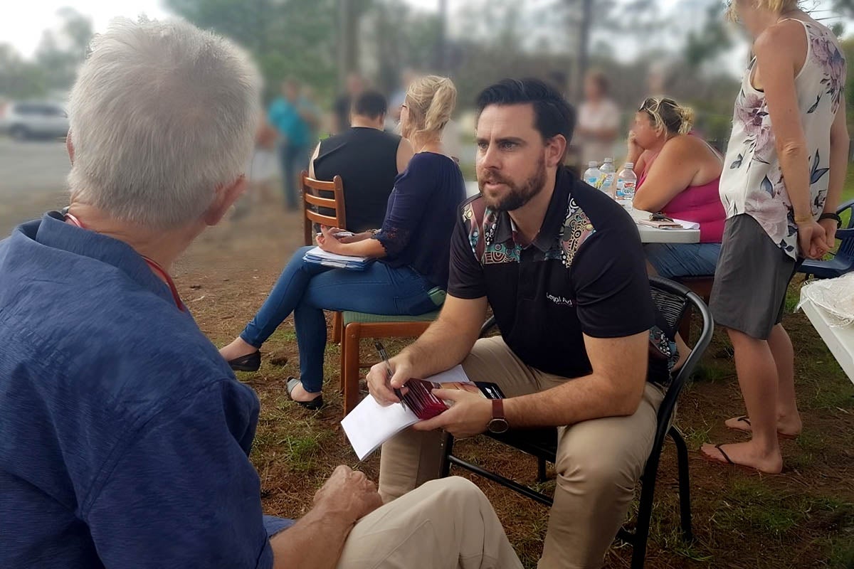 A lawyer sits in a chair outside, leaning forwards. He is speaking to another man and holding some brochures.