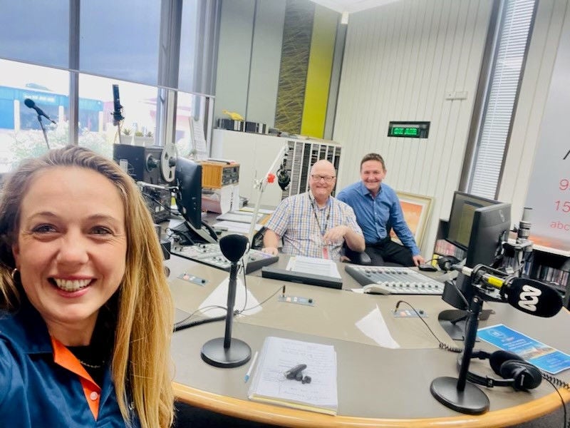 A woman smiles in the foreground of a radio studio as she takes a selfie of herself and two others who are smiling across the desk from her. On the desk is stuff associated witha radio studio such as microphones, headphones and mixing desks. 