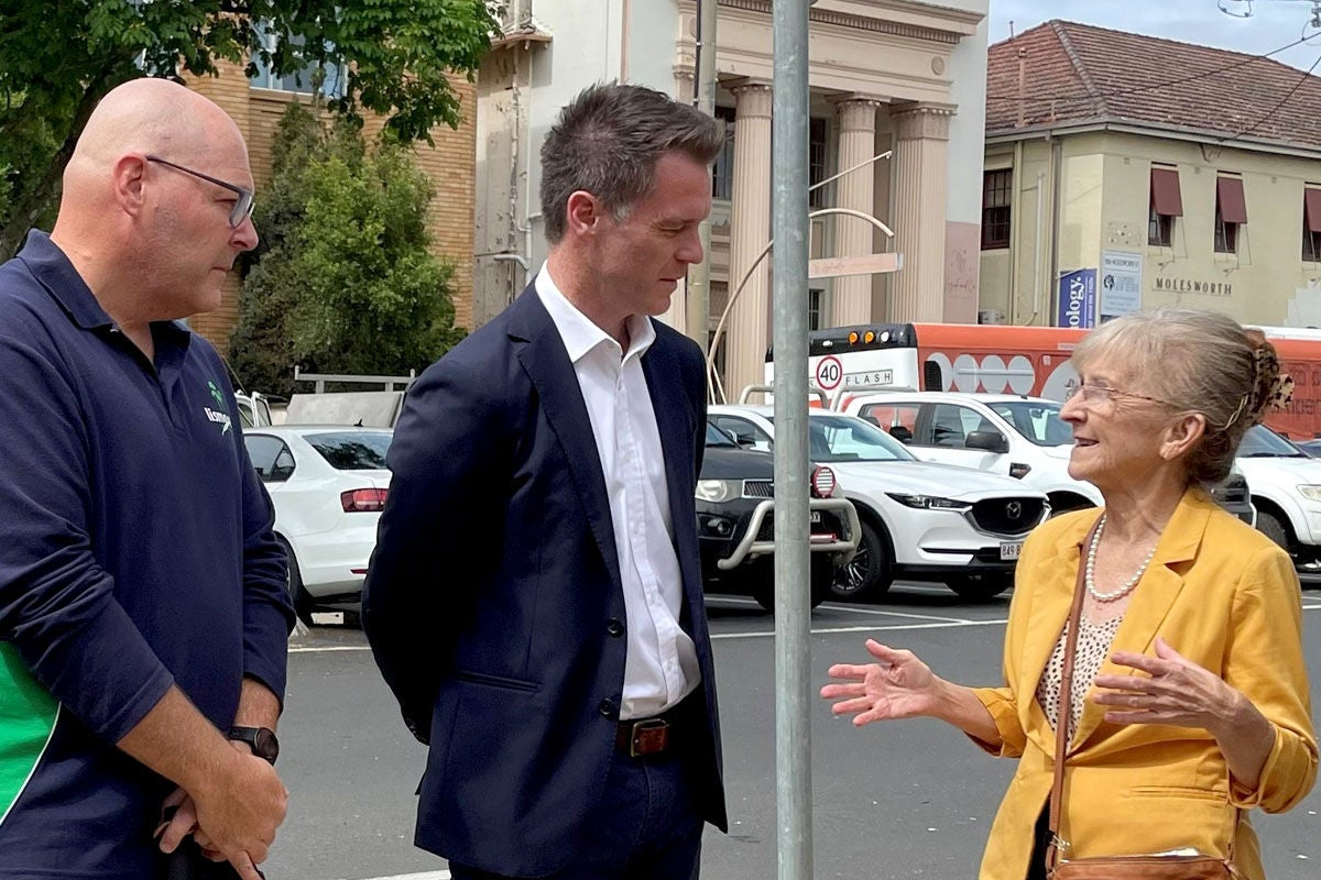 The mayor of Lismore Steve Kreig stands next to Chris Minns and Janelle Saffin. Janelle and Chris are engaged in conversation. They are all standing on a street, parked cars are visible behind them.