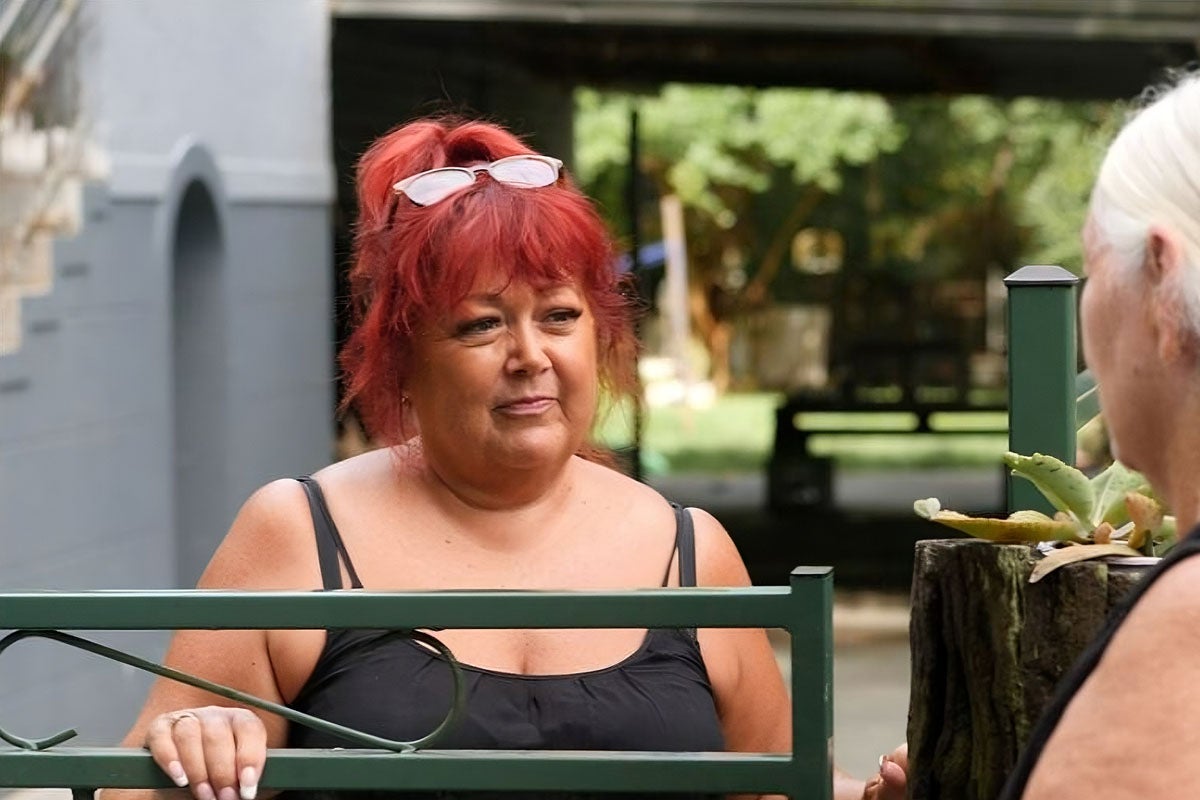 A woman with bright red hair stands behind a gate. She is smiling at another woman who has her back to camera.