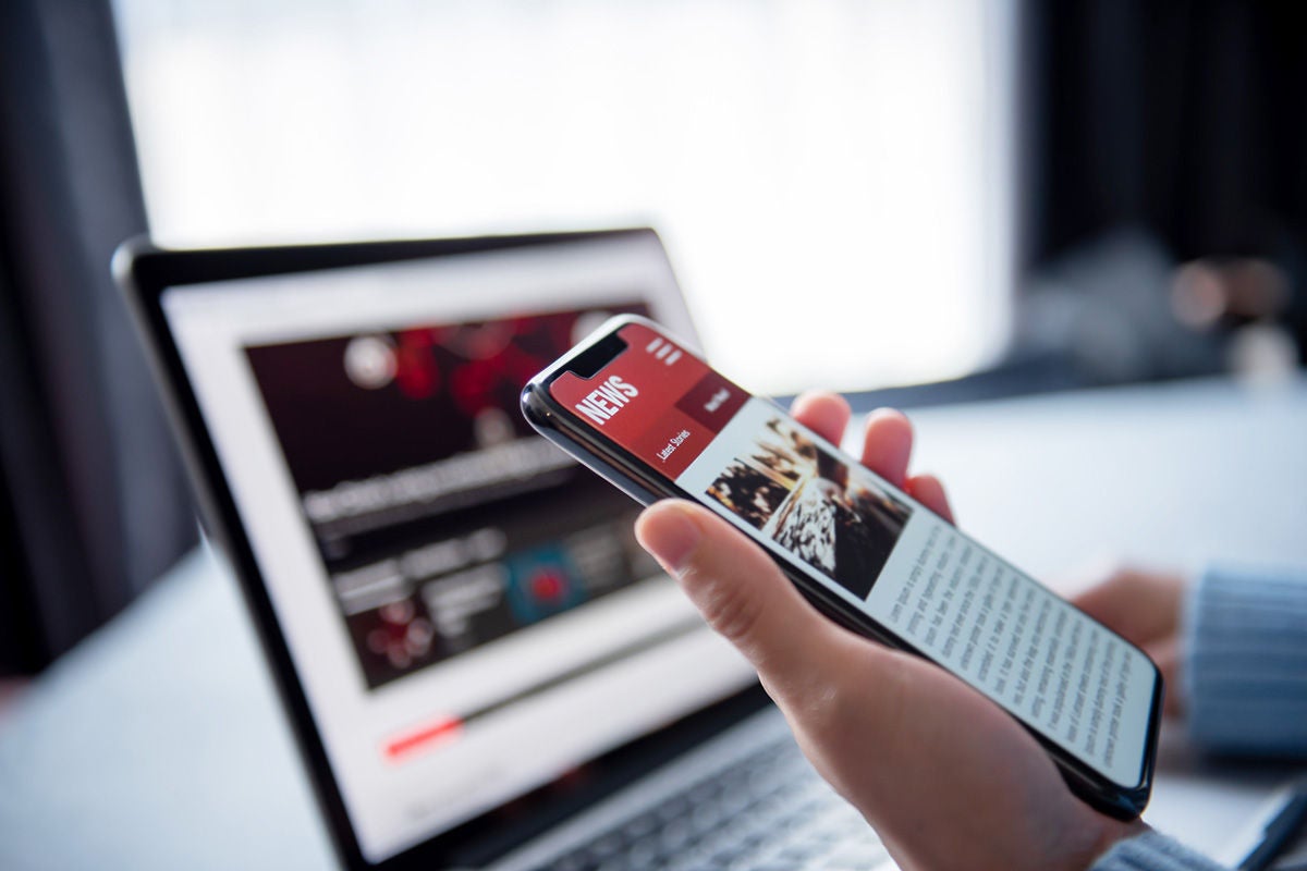 A hand holds a phone open to a news screen. Behind the phone an open laptop can be seen, but the screen is blurry.