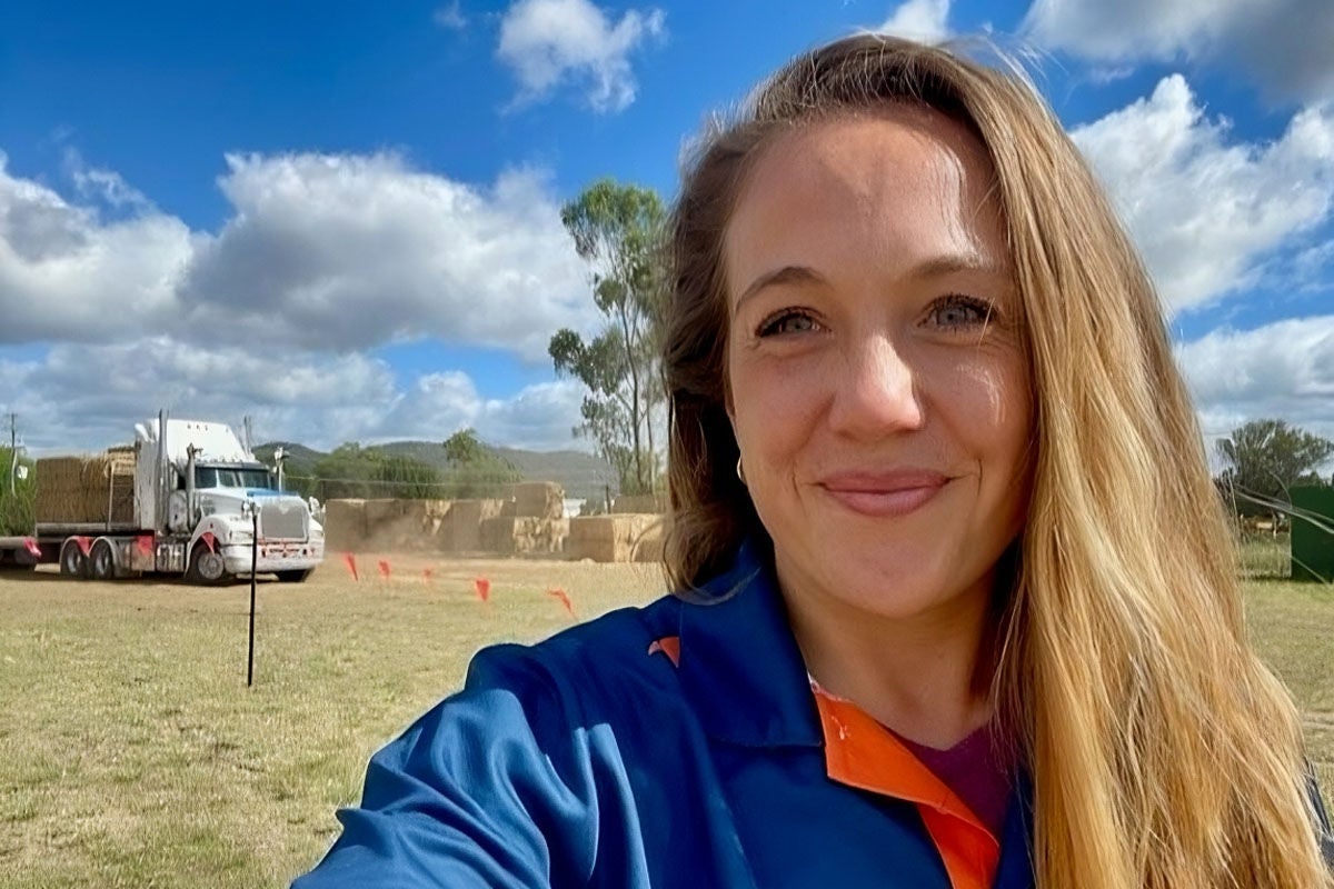 A woman in a blue and orange shirt takes a selfie from an open paddock. Behind her a truck appears to be delivering hay bales