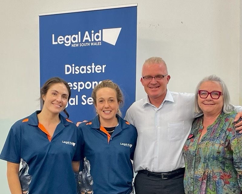 Mel, Liz, Mal and Donna stand smiling in front of a Legal Aid NSW Disaster Response Legal Service banner